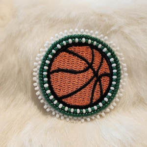 May include: A round, beaded pin featuring a basketball design. The basketball is orange with black lines, surrounded by a green beaded border, a white beaded edge, and a white fluffy background.