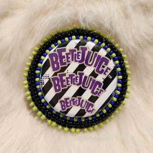 May include: A round pin with the text "Beetlejuice" in purple and white, set against a black and white striped background. The pin is surrounded by black, blue, and green beads.