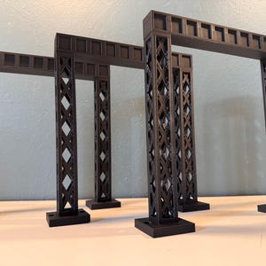 以下が含まれることがあります： 黒い建築様式のアーチモデルが3つ。各モデルは長方形のベース、ダイヤモンド格子模様の垂直サポート、長方形の切り欠きのある水平な上部を備えています。モデルは白い表面に配置されています。