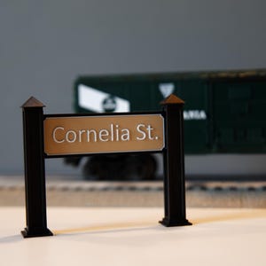 May include: A miniature street sign with the text "Cornelia St." in a serif font. The sign is tan with black posts and a decorative top. A dark green train car is in the background.