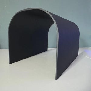 May include: A black, arch-shaped object with a gray trim. The object is made of a rigid material and stands upright on a white surface. The shape resembles a small tunnel or a curved shelter.