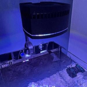 Könnte beinhalten: Eine schwarze Aquarienbeleuchtung hängt über einem Glasaquarium, das mit Wasser gefüllt ist. Das Licht hat eine rechteckige Form mit Schlitzen und einem dünnen, beleuchteten Streifen. Das Wasser im Aquarium reflektiert blaues Licht.