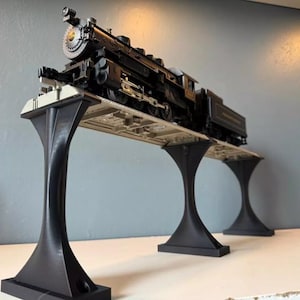 May include: A detailed model train, predominantly black, rests on a bridge-like structure with three black supports. The train features intricate detailing, including a prominent smokestack and various mechanical components. The bridge is supported by three black, stylized pillars.