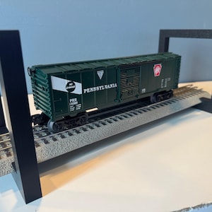 May include: A dark green Pennsylvania Railroad boxcar model with the text "Pennsylvania" and a red logo. The model train car sits on a grey track and is supported by a black frame. The model is a miniature replica of a freight car.