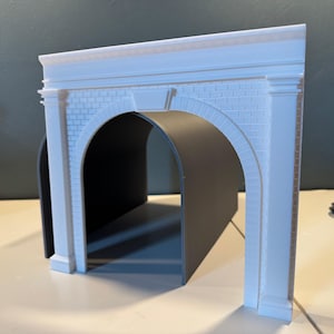 May include: A white model train tunnel with a brick facade and arched openings. The tunnel has a rectangular shape with a flat top and is designed to simulate a bridge or tunnel for model train layouts. The interior of the tunnel is black.