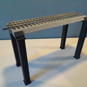 以下が含まれることがあります： グレーのテクスチャードデッキと黒いサポート柱を備えた模型鉄道の橋。橋は、銀色のレールを備えた2つの平行なトラックをサポートしています。 模型鉄道レイアウト用に設計されています。