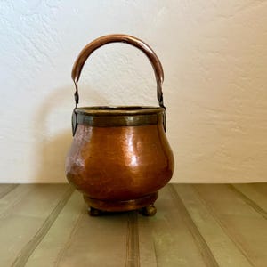 May include: A small, round, hammered copper pot with a polished finish, featuring a curved metal handle attached to the sides and three small, rounded feet at the base. The rim of the pot is accented with a darker, metallic band.