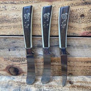 May include: Set of three butter knives with brown and white handles. The handles have a floral design. The blades are stainless steel.