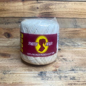 Könnte beinhalten: Eine Spule aus cremefarbenem Baumwollgarn, verpackt in durchsichtigem Kunststoff. Das Etikett trägt die Aufschrift "SOUTH MAID" mit einer Silhouette eines Frauenprofils und "100% MERCERIZED COTTON" in weißer Schrift auf bordeauxrotem Hintergrund. Das Garn liegt auf einer Holzoberfläche.