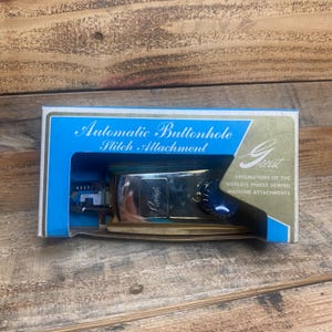 Puede incluir: Un accesorio de puntada de ojal automático de Greist, en su caja original. La caja es azul y blanca con detalles dorados. El accesorio es plateado y dorado con una perilla azul. La caja dice "Originators of the World's Finest Sewing Machine Attachments."