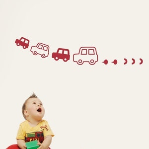 Può includere: Un adesivo da parete con una fila di quattro auto bordeaux stilizzate di varie dimensioni, disposte in un leggero arco verso l'alto. L'auto più grande sulla destra è seguita da una serie di piccole forme bordeaux di dimensioni decrescenti.