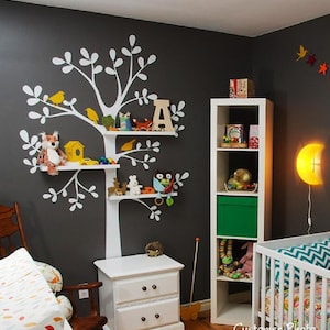 Könnte beinhalten: Kinderzimmer mit grauer Wand mit einem weißen baumförmigen Regal mit Spielzeug und Dekorationen. Ein weißer Nachttisch und ein Bücherregal sind ebenfalls vorhanden. Ein Kinderbett und eine mondförmige Wandleuchte vervollständigen die Raumdekoration.