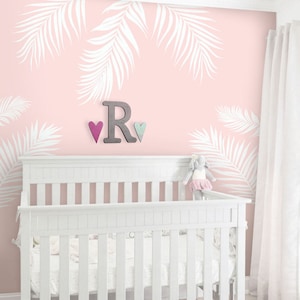 Peut inclure: Un lit b&eacute;b&eacute; blanc avec un mur rose orn&eacute; d'un motif de feuilles de palmier blanches. Le mur pr&eacute;sente &eacute;galement une lettre R en bois et deux petits c&oelig;urs.