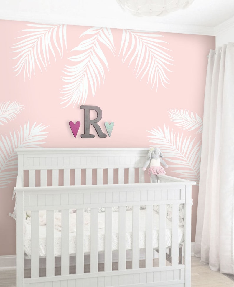Peut inclure: Un lit b&eacute;b&eacute; blanc avec un mur rose orn&eacute; d'un motif de feuilles de palmier blanches. Le mur pr&eacute;sente &eacute;galement une lettre R en bois et deux petits c&oelig;urs.