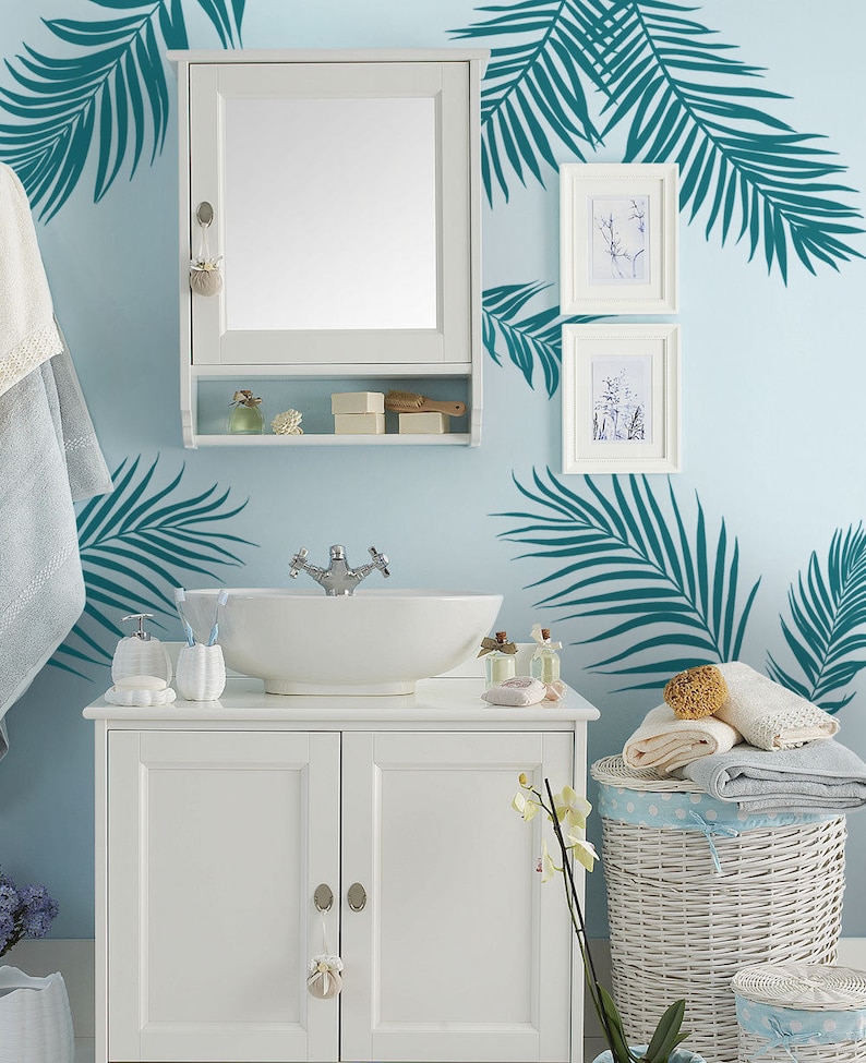 Peut inclure: Vasque de salle de bain blanc avec un lavabo en porcelaine blanche et un robinet chrom&eacute;. Le meuble sous vasque a deux portes avec des boutons argent&eacute;s. Un panier &agrave; linge en osier blanc avec un motif &agrave; pois bleus et blancs se trouve &agrave; droite du meuble sous vasque. Le mur derri&egrave;re le meuble sous vasque est bleu clair et comporte des autocollants en forme de feuilles de palmier turquoise.
