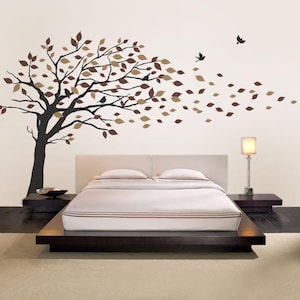 May include: A wall decal featuring a stylized tree with brown and tan leaves blowing in the wind. The tree is black, and birds are flying nearby. The decal is above a bed with a white headboard and a wooden frame.