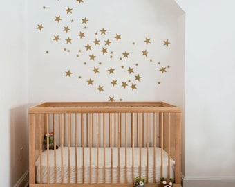 May include: A wooden crib with a mattress and a few stuffed animals. The wall behind the crib is decorated with gold star decals. The crib is made of light-colored wood and has vertical slats.