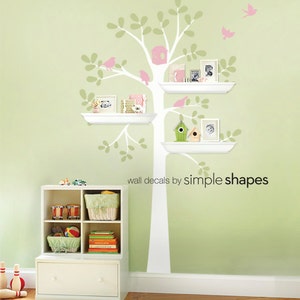Pode incluir: Um decalque de parede com uma &aacute;rvore branca com prateleiras, adornada com p&aacute;ssaros rosa e uma casinha de p&aacute;ssaros. As prateleiras cont&ecirc;m fotos emolduradas e itens decorativos. O texto "wall decals by simple shapes" &eacute; vis&iacute;vel.