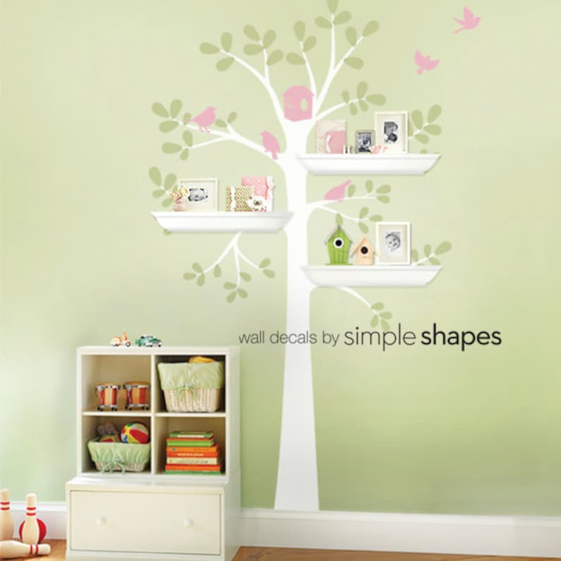 Pode incluir: Um decalque de parede com uma &aacute;rvore branca com prateleiras, adornada com p&aacute;ssaros rosa e uma casinha de p&aacute;ssaros. As prateleiras cont&ecirc;m fotos emolduradas e itens decorativos. O texto "wall decals by simple shapes" &eacute; vis&iacute;vel.