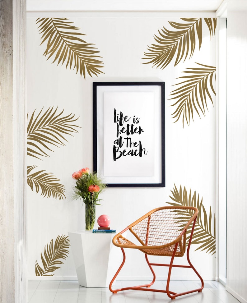Peut inclure: Un mur blanc avec des autocollants en forme de feuilles de palmier dor&eacute;es, une impression encadr&eacute;e avec le texte "La vie est meilleure &agrave; la plage", une table d'appoint blanche avec un vase de fleurs roses et une chaise en osier avec un cadre en m&eacute;tal orange.