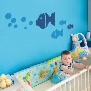 Può includere: Un set di adesivi murali in vinile blu scuro raffiguranti un grande pesce che soffia bolle, accompagnato da tre pesci più piccoli. Gli adesivi sono applicati su una parete azzurra sopra una culla bianca con un paracolpi colorato.