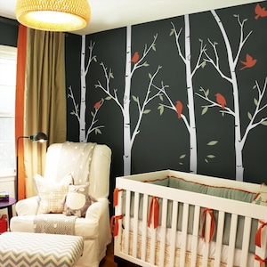 May include: A nursery with a white crib, a white armchair, and a patterned ottoman. The dark gray wall features white birch tree decals with red birds and green leaves. A woven light fixture hangs from the ceiling.