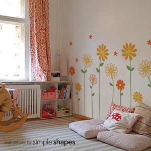 May include: A white wall with orange and yellow flower wall decals. The decals are arranged in a vertical line and feature green stems. There are two pink and white patterned cushions on the floor in front of the wall.