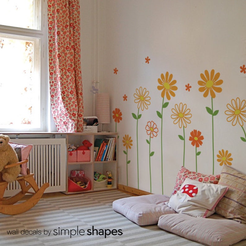 May include: A white wall with orange and yellow flower wall decals. The decals are arranged in a vertical line and feature green stems. There are two pink and white patterned cushions on the floor in front of the wall.