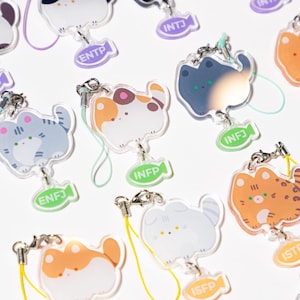 May include: A collection of clear acrylic cat charms with various color schemes and personality type labels. Each charm has a silver clasp and a colored lanyard. The cat designs include black, white, orange, and gray, with labels like ENTP and INFP.
