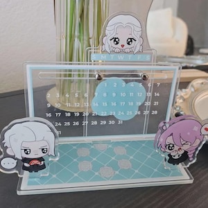 May include: A clear acrylic calendar with a light blue patterned base and a calendar grid. The calendar features cartoon character accents. The top of the calendar has the letters MTWTFS. The calendar is decorated with cartoon character accents.