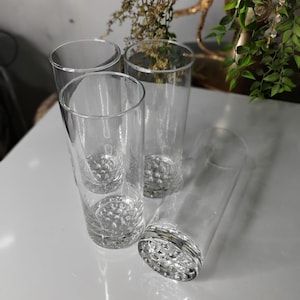 May include: Four clear, cylindrical drinking glasses. The bases of the glasses have a textured, bubble-like design. The glasses are arranged on a white surface, with some greenery visible in the background.