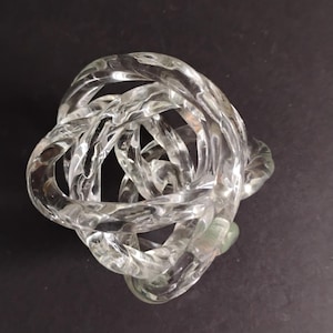 May include: Clear glass decorative knot sculpture. The sculpture is made of intertwined glass loops, creating a complex, knotted design. The glass has a slight green tint in some areas, and the background is black.