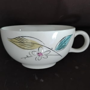 May include: White ceramic teacup with a floral design. The cup features a handle and is decorated with a stylized flower and leaves in yellow, blue, and black. The cup has a round base.