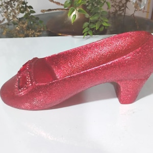 Puede incluir: Un zapato rojo brillante con un tacón pequeño y una hebilla decorativa. El zapato es de un tono rojo vibrante y parece estar hecho de un material brillante. El zapato es un objeto decorativo.