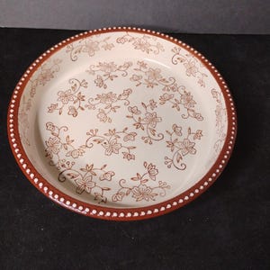 May include: Round, off-white ceramic pie dish with a brown floral pattern. The rim is brown with white dots. The dish has a classic, vintage aesthetic, suitable for baking and serving.