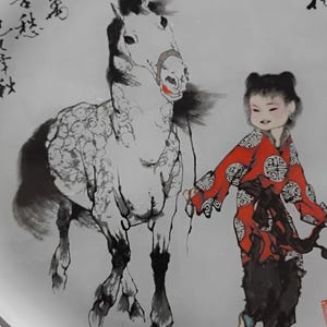 May include: A decorative plate featuring a traditional Chinese painting. The artwork depicts a white horse with a black mane and legs, led by a person in a red robe. The plate has calligraphy around the edge.