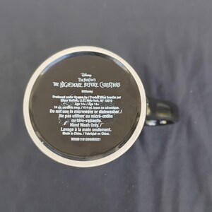 May include: Black ceramic mug with text from Tim Burton's "The Nightmare Before Christmas". The mug has a black handle and a white rim. The base of the mug includes product information and care instructions.