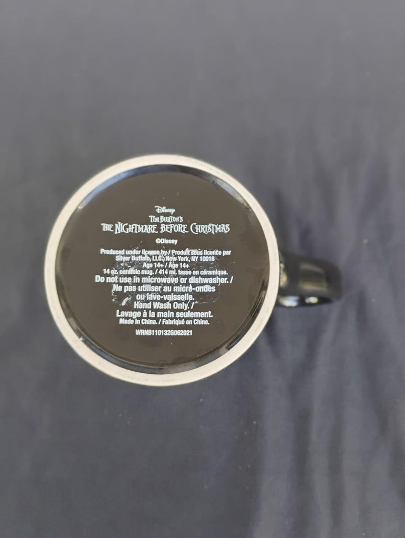 May include: Black ceramic mug with text from Tim Burton's "The Nightmare Before Christmas". The mug has a black handle and a white rim. The base of the mug includes product information and care instructions.