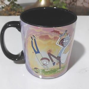 May include: A ceramic mug with a black handle and interior. The exterior features a colorful cartoon scene with two characters in a dynamic pose against a sunset-like background. The mug is designed for beverages.