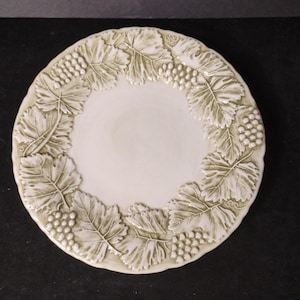 May include: Off-white ceramic plate with a raised grape and leaf design around the rim. The grapes and leaves are a muted green color. The plate has a slightly scalloped edge and a smooth, flat center.