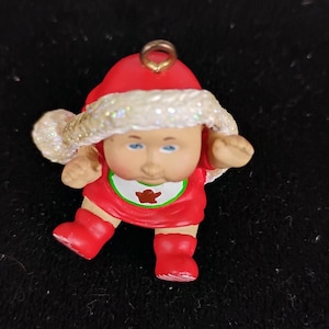 May include: A Christmas ornament featuring a baby doll figure in a red Santa suit and hat with a glittery white trim. The doll has blue eyes and a bib with a brown bear graphic. The ornament has a gold loop for hanging.