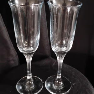 May include: A pair of clear glass champagne flutes featuring tall, slender bowls and stems with vertical ribbed detailing at the base of the bowls and along the stems, leading down to circular, flat bases for stability and balance.