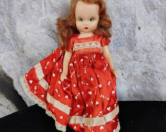 May include: A vintage doll with red hair and a red dress adorned with white hearts. The dress features white lace trim and a cream-colored ribbon detail. The doll has green eyes and red lipstick.