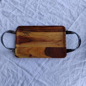 May include: A rectangular wooden serving tray with two black leather handles. The tray is made of acacia wood and has a natural wood finish. The tray has the text "HANDCRAFTED IN INDIA" and "EACH PIECE IS UNIQUE" engraved on the surface.