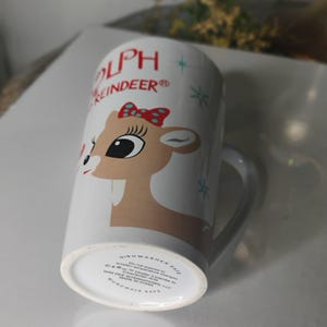 May include: White ceramic mug featuring a cartoon illustration of Rudolph the Reindeer with a red bow. The mug has the words "Rudolph the Reindeer" in red and blue. The mug is dishwasher and microwave safe.