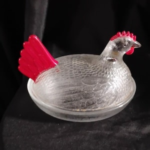 May include: A clear glass hen-shaped dish with a red comb and tail. The hen is sitting in a round dish, and the body has a textured pattern. The comb and tail are a bright red color.