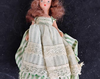 May include: A vintage doll with brown hair and red lips. The doll wears a green and white striped dress with lace trim. The dress has a ruffled hem. The doll's arms are at its sides.