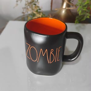 May include: Black ceramic mug with an orange interior. The word "ZOMBIE" is written in orange on the front of the mug. The mug has a curved handle and is sitting on a white surface.