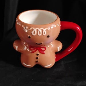 May include: A ceramic gingerbread man mug with a red handle and bow tie. The mug is brown with white icing details and a white interior. The mug is designed to look like a gingerbread man.