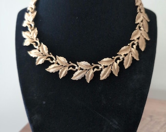 May include: A gold-toned necklace featuring a leaf design. The necklace is displayed on a black mannequin. The leaves are detailed and arranged in a repeating pattern, creating a classic and elegant look. The necklace is a statement piece.
